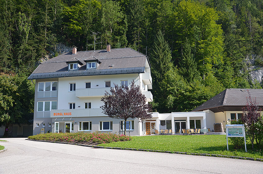 Three storey white house with annex and a terrace in a park area with woods in the background.