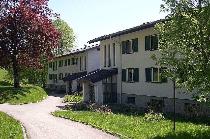 Long stretched two storey building with green window shutters in a park area, a road leading to the entrance and a tree with red leaves on the side.